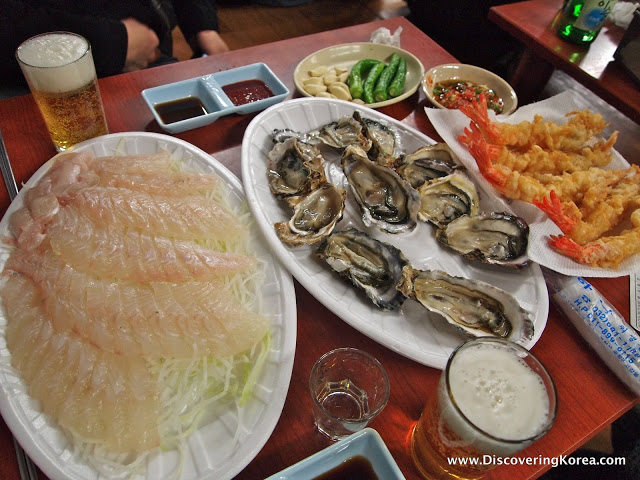 A seafood platter, on a wooden table. On the left is fresh sashimi, white fish on a white dish, in the center are freshly shucked oysters, and on the right deep fried battered prawns. Condiments and two glasses of beer behind.