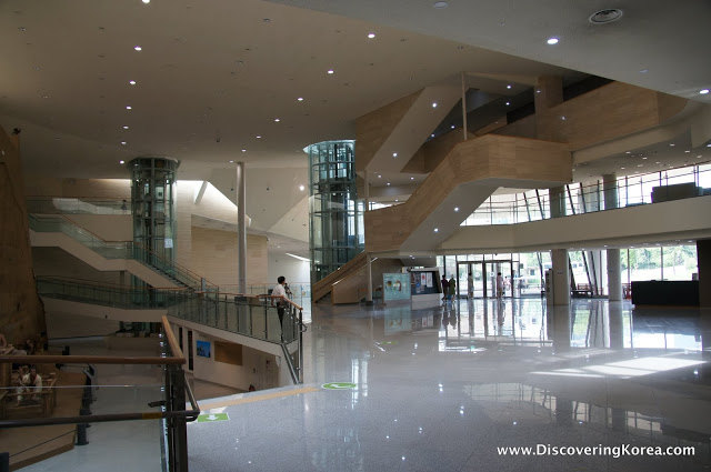 Interior of the Seoul Baekje Museum, a light filled atrium with spotlights in the ceiling, glass walls and a light colored polished floor.