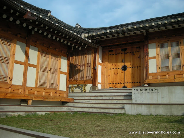 L-shaped wooden house in Bukchon hanok village, with grass in the front, a verandah and steps up to the front door. Shutters cover the windows.