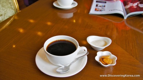 A wooden table with a white cup containing black coffee, a spoon, and a bowl of sugar and a small bowl of cookies.