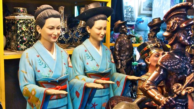 A close up of two Korean statues, ladies with their hair in a bun, wearing blue kimonos, embroidered with red flowers and green leaves, standing with their hands out, palms facing upward. Behind are various statues and vases at the Seoul Folk Flea Market.