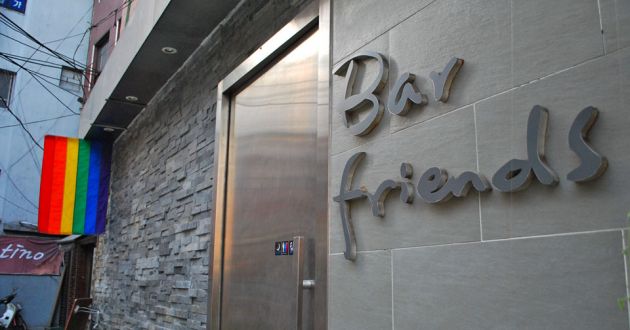 The entrance to a bar with a sign on the right of the frame saying Bar Friends on stone, a metal door and slate wall, with a rainbow flag hanging from the eaves to the left of the frame.
