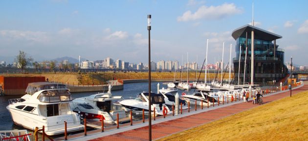 A line of boats moored at Seoul Marina, with a red brick walkway and grass on the river shores. In the background is the large glass marina building and soft focus cityscape against a blue sky.