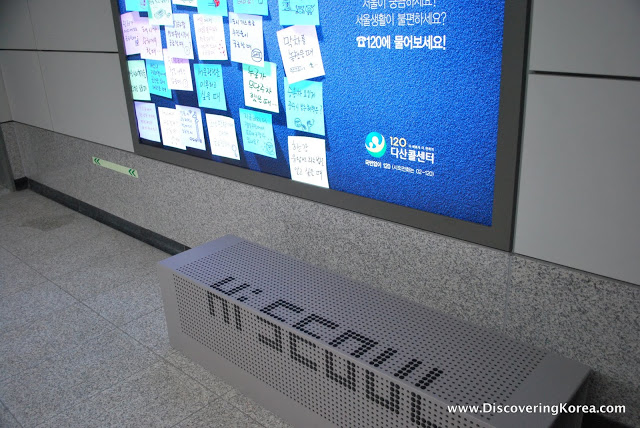 A gray metal bench with ventilation holes and the words 'Hi Seoul' in black lettering. To the top of the frame is a blue sign with post it notes in Korean.