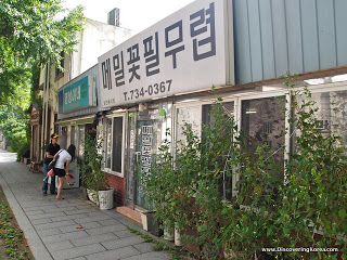 View of shopfronts of noodle bars in Seoul. to the right of the frame is glass doors with trees outside, to the left is the sidewalk and two people looking through a window.