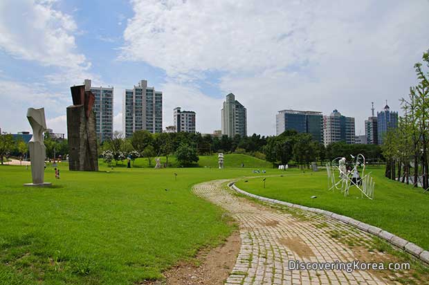 Seoul's Olympic Park, a brick walkway with lawns on either side, large geometric sculptures to the left of the frame, and tall buildings in the background, with a blue, cloudy sky.