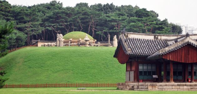 The entrance to Seoul Royal Tombs, with a wooden building to the right of the frame, with a traditional curved roof, to the center of the frame is a large round mound of grass surrounded by a stone wall, in the foreground are neat lawns and in the background a pine forest.