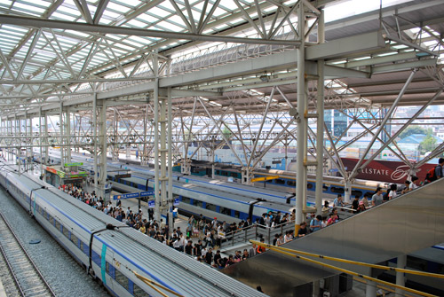 Looking down on trains at Seoul station, large groups of pedestrians on the platforms, white metal roof structure with a glass ceiling.