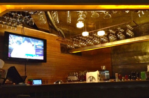 A bar, with a television on the wall, glasses hanging from shelves above, dimly lit.