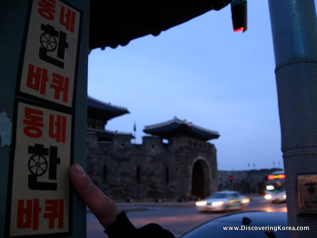 A finger points at sticker on a lamp post, with soft focus buildings and cars in the background, at dusk.