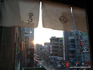 Street scene with fabric hanging from the top of the frame, looking out onto evening sunshine with buildings, cars and pedestrians.