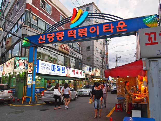 A street in Seoul, with a blue and red sign over the street, in the background are lots of shopfronts and restaurants and people walking around.