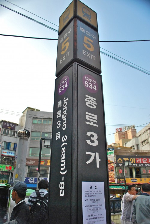 A subway sign in Seoul, a black pillar with Korean writing and buildings and pedestrians behind, with a blue sky.