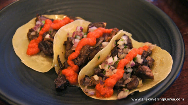 Three soft tacos with meat, salsa and a red sauce filling, on a black plate in Seoul.