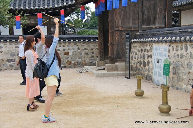 People standing in a line in a stone courtyard at Namsangol throwing sticks at a target. A stone building behind, with dark wooden doors.