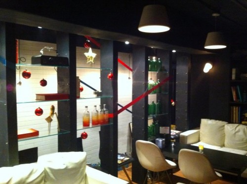 Interior of a bar, with black and glass cabinetry, cream and black furniture, red and green bottles in the background.