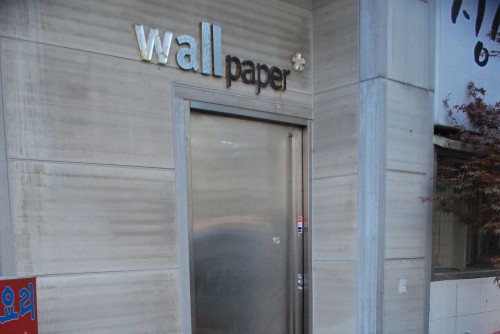 View of an entrance to a bar, a stone block building with a metal door and a sign above it saying wall paper in metallic lettering.