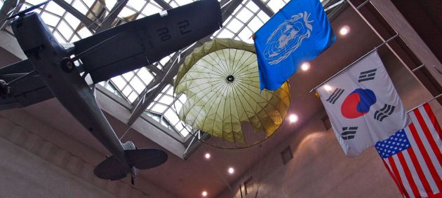 Looking upwards towards a skylight ceiling in the background, is a war airplane hanging from the ceiling, with flags to the right of the frame and an open parachute in the center.