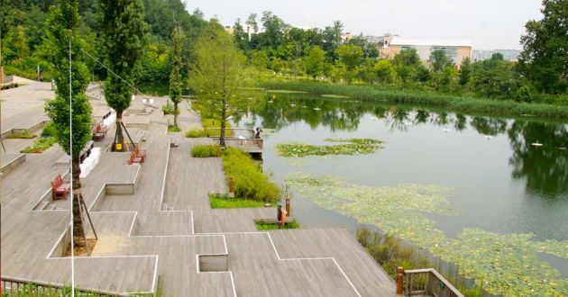 Geometric steps with bench seating, ornamental trees, leading down to the lake at West Seoul Lake Park. The clear water of the lake reflects the trees and grasses in the background that surround it.