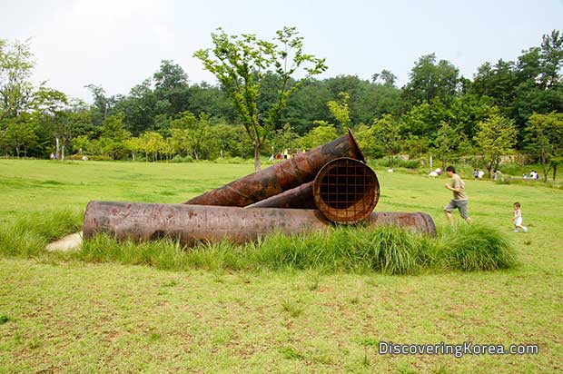 Three large metal rusting pipes in the middle of a grass lawn with trees and shrubs in the background and a man and a child running in the background.