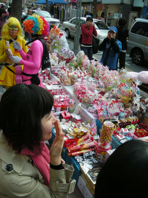 A table, laden with candy, in the street, for White Day in Korea. Two colorfully dressed people with multicolored wigs on the left of the frame, and other pedestrians around the table.