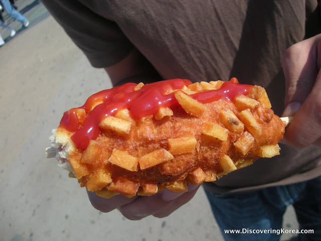 Close up of a man holding a large deep-fried corndog with tomato ketchup on top.