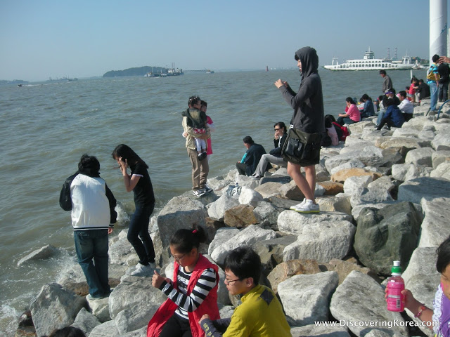 People on a rocky bulkhead at Wolmido island, taking photographs. On the left of the frame is the sea, showing a ferry docking at the port.