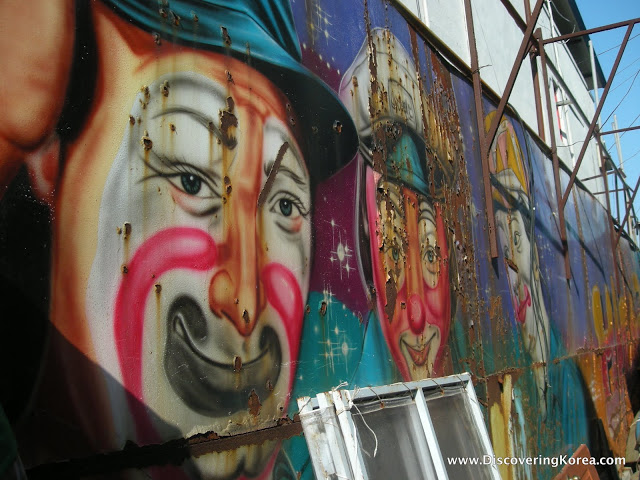Multicolored mural of a clown face on a wall in Wolmido Island. Scaffolding to the right of the frame.