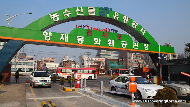 Entrance to Yangjae Flower Market, showing a green archway over the road, with Korean lettering, cars passing through barriers underneath and shop fronts in the background with a blue sky.