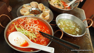 A large metal dish with noodles in a red sauce, behind this is a metal bowl with clear broth soup and steamed dumplings.