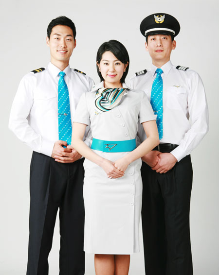 Three airline staff, two men dressed in white shirts, black pants and bright blue ties, one is wearing a Captain's hat. In the middle is a woman in a white dress with a bright blue sash and striped scarf.