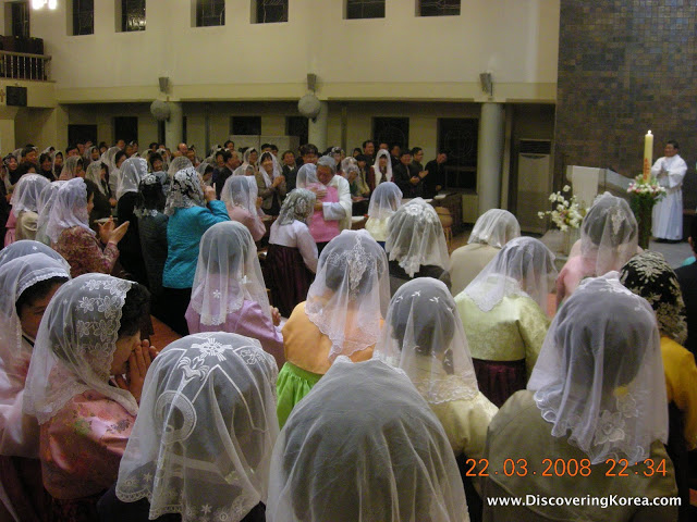 A group of worshippers with white veils covering their heads and a pastor dressed in white robes at the front of the congregation with a large candle.