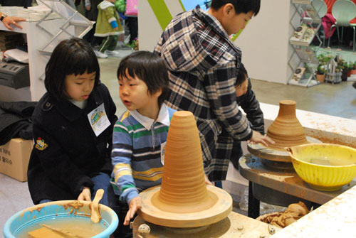 Three children making pottery with clay. There are two pottery wheels and a plastic blue bucket with fresh clay. The background is of a pottery shop.