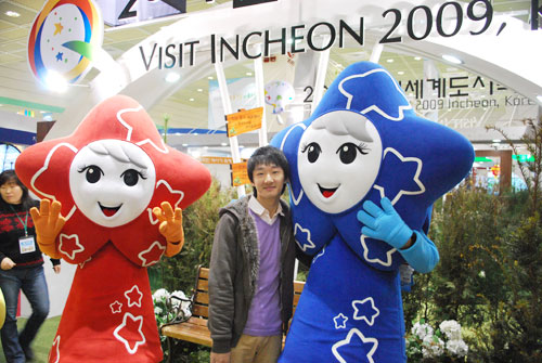 Two people wearing costumes featuring stars as heads, one is blue one is red. In the background is a man standing in front of a big glass sign, with plants and a bench underneath it.