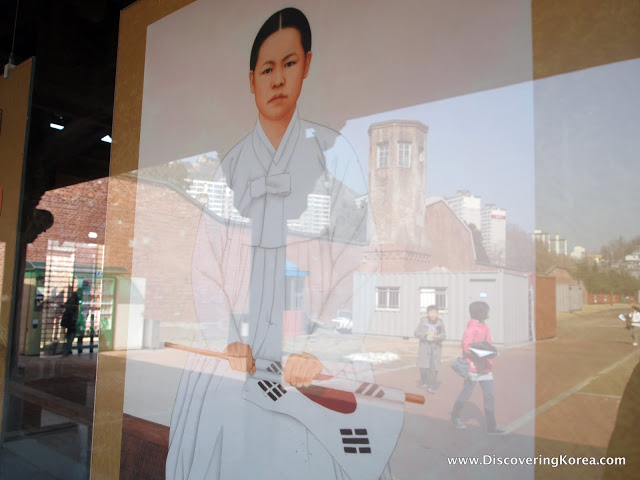 Pictured through glass, a painting of a person in white robes, with hands clenched around a Korean flag.