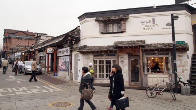 A street view of Samcheong dong neighborhood in Seoul. A pedestrian area with people walking, with the main street to the left of the frame. In the background is a white and gray building with a bicycle parked outside it, and various shop fronts extending down the main street.