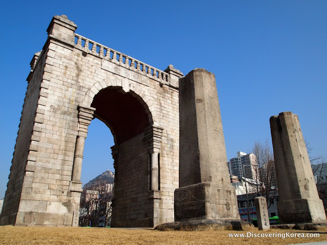 A large stone archway with two concrete pillars in front of it, in the background is modern Seoul, with a blue sky in bright sunlight.