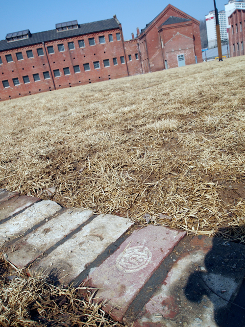 A vertical image with the Seodaemun prison building in the background. Red brick and imposing with small dark windows. In the foreground is dried grass and a brick pathway. One of the bricks in the path has a symbol carved into it.