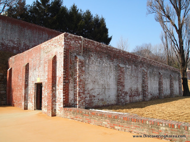 An imposing brick structure at Seodaemun prison. The square building is red brick, with white discoloration. In the foreground is a clay courtyard with a low brick wall, and in the background is blue sky and trees on a bright sunny day.