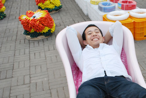 A man wearing a pink striped shirt and jeans reclines in a pink chair with colorful plastic boxes and fake flowers behind. The background is a concrete brick surface.