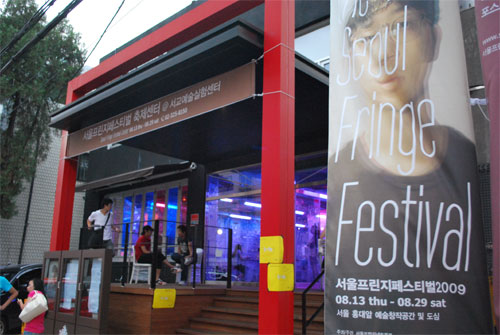 A building with brick steps leading up to the brightly lit glass entrance of the Seoul Fringe Festival art space. On the right of the frame is a vertical banner advertising the festival.