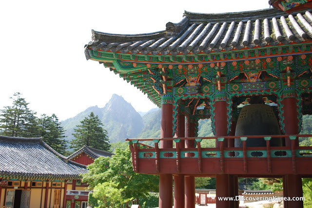The bell tower of a Buddhist temple with curved roof and ornate turquoise painting and carving in the eaves. In the background are further buildings and views over the forest and mountains.