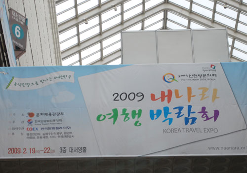A large billboard sign for the Korean Travel Expo in 2009. Text in various colors on a light blue background. A glass roof can be seen in the background.