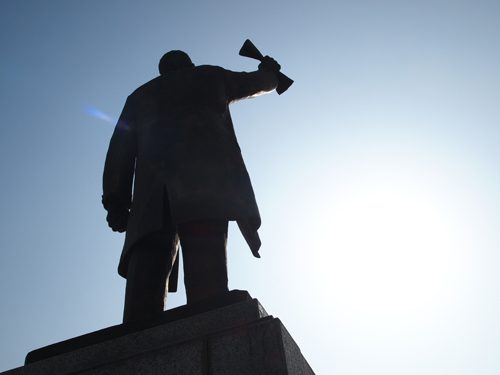 A close up of a statue of an independence activist, his right arm up holding a rolled up paper. The background is blue sky and bright sunshine.