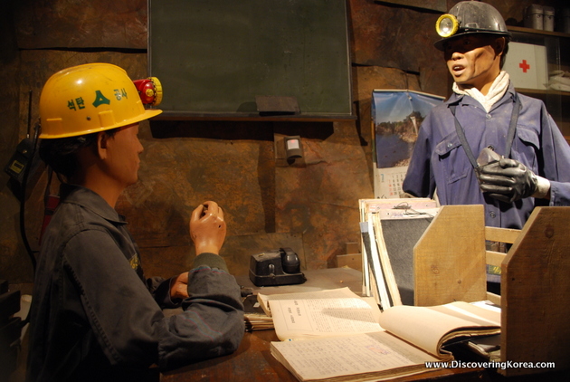 Models in the Taebaek coal museum. The scene depicts two people in an office one behind the desk wearing a yellow hard hat with a light, and a man standing the other side of the desk with blue overalls and a mining lamp.