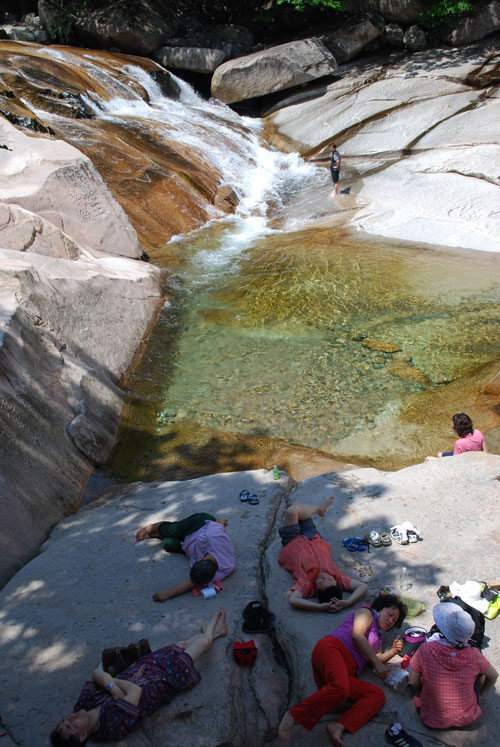 A vertical image of a waterfall emptying into a shallow pool between large flat rocks. People lying down in the shade on a sunny day.