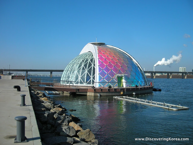 A glass dome floating stage on Yeouido island, on the water with the city behind it.